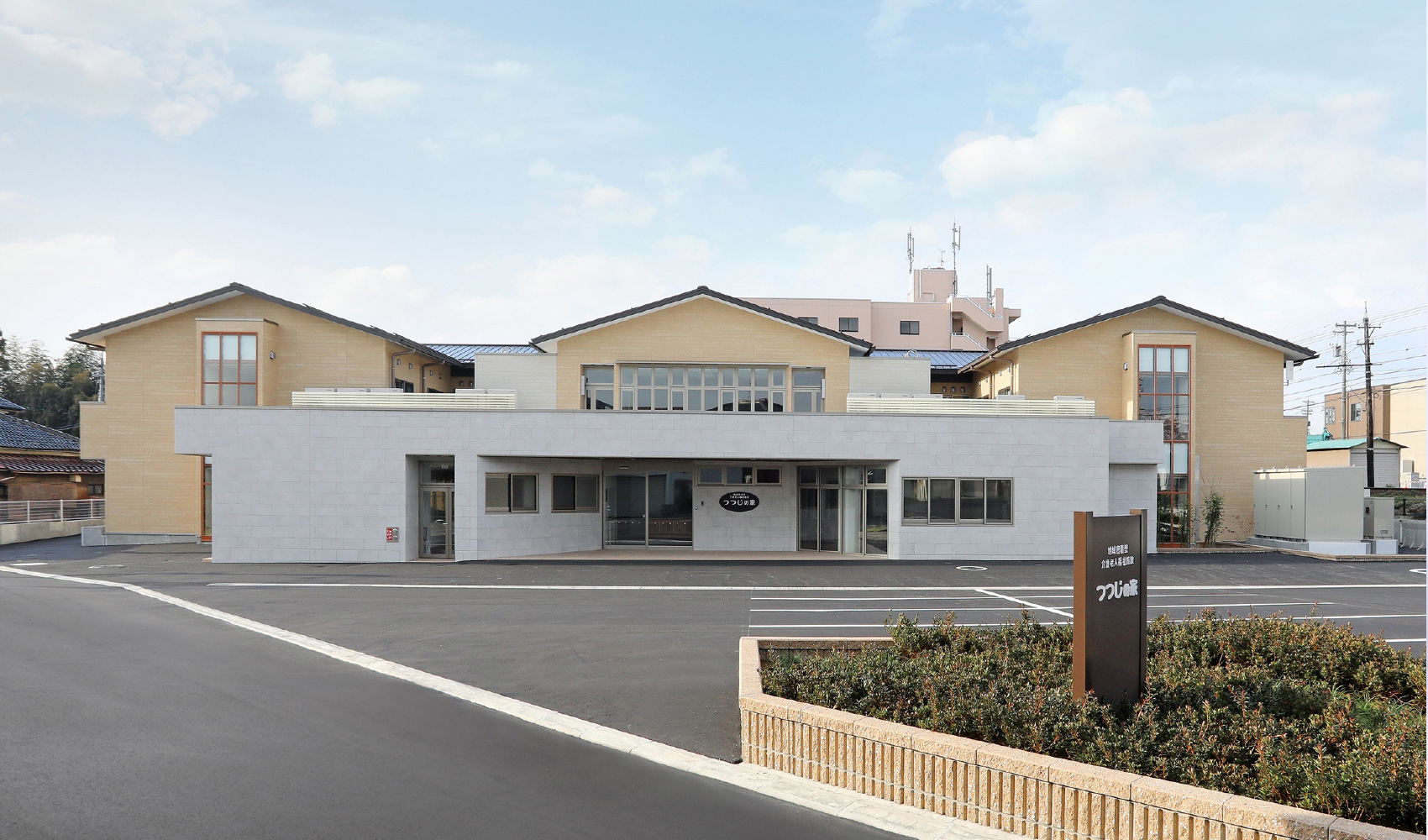介護施設の建物外観。白とベージュを基調とした現代的な2階建て施設。正面駐車場から撮影。